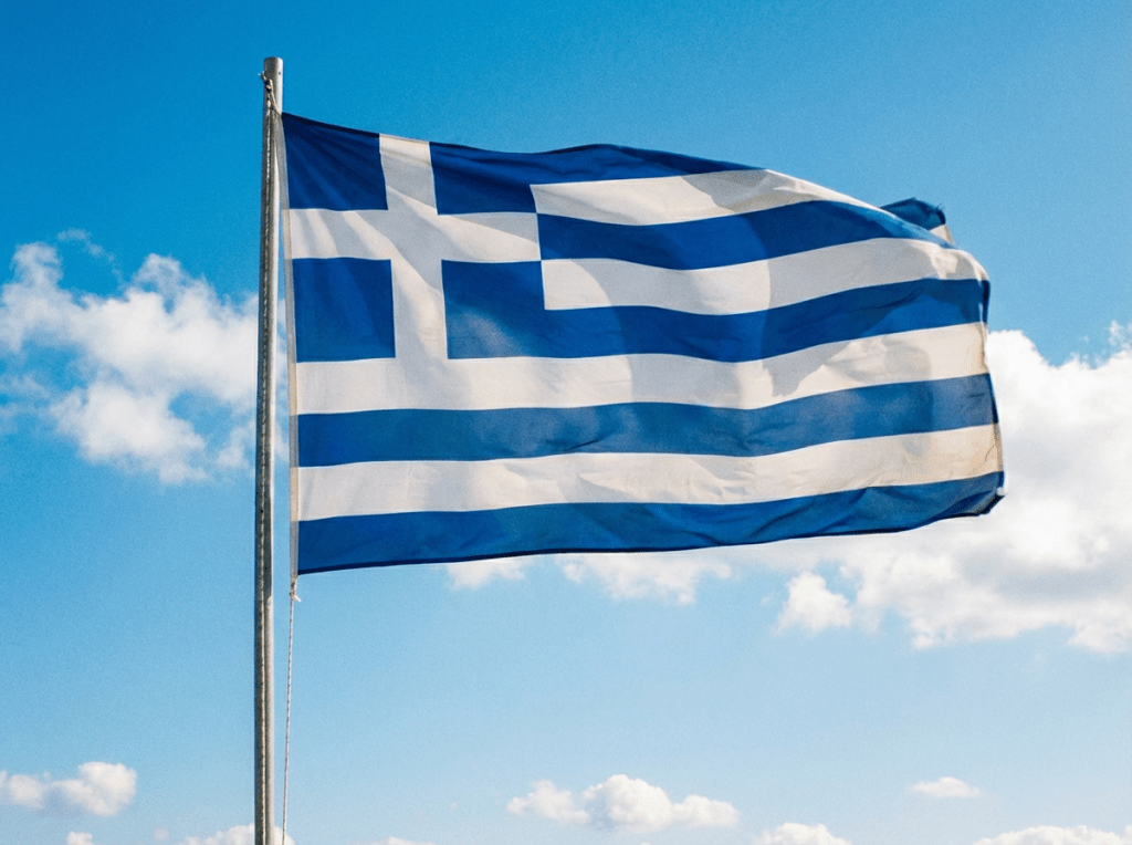 A Greek flag waving on a flagpole overlooking a white chapel and the sea.